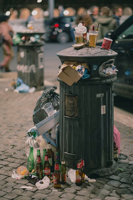 A black, cylindrical public trash bin with a domed lid is situated on a cobblestone pavement in an urban outdoor setting. The top of the bin is covered with various discarded items, including used paper cups containing beverages with ice and straw, a crumpled snack wrapper, and other small pieces of rubbish. The front of the bin has a slot for paper and a small opening for waste, with several used bottles, glass and plastic, placed at its base, some on the ground and others leaning against the bin. There are also multiple crumpled tissues and packaging materials scattered around the base, indicating recent improper disposal. In the background, a grey bin with the label 'Rubbish' is visible, along with blurred figures of people walking and cars parked along the street, suggesting a busy city environment. The scene reflects typical urban waste accumulation that could be managed through private rubbish clearance services, such as those offered by Rubbish Clearance Croydon, to prevent littering and waste overflow in public areas.