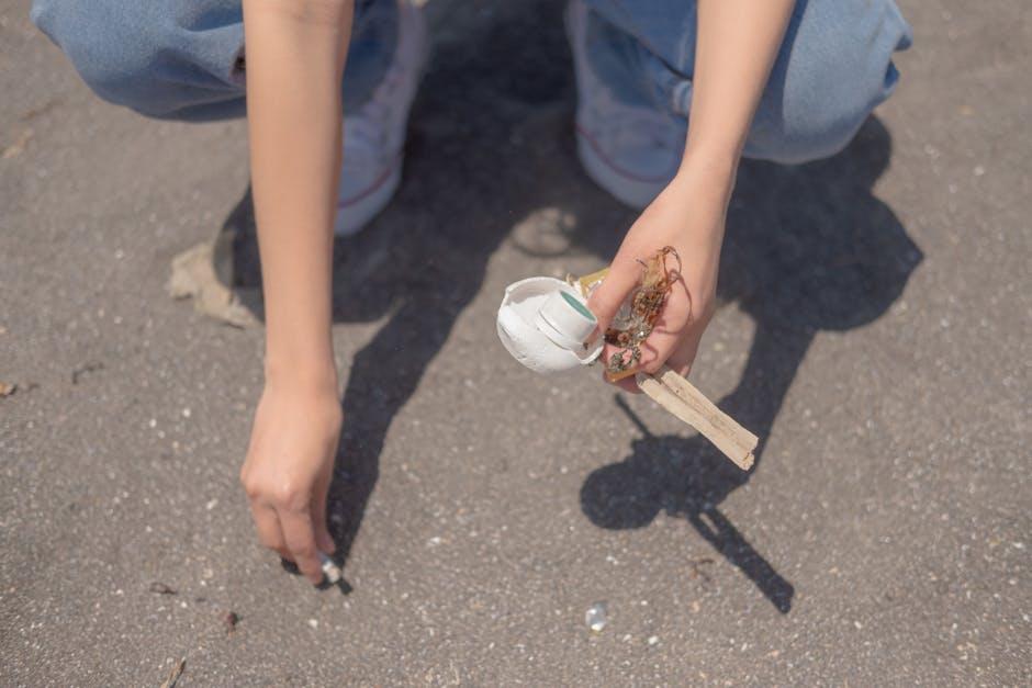 A person crouching on a paved outdoor surface, visible from the knees down, wearing light blue jeans and white sneakers. The individual is holding a small, broken wooden object with a white ceramic or plaster ball attached, which appears to be a damaged decorative item or craft. The person's right hand holds the item, while the left hand rests on the ground with fingers slightly curled. The background shows the smooth asphalt surface with small pebbles and debris, and a shadow cast by the person can be seen to the right. This scene suggests an individual engaging in waste collection or disposal of small household or craft debris, which could relate to a rubbish removal service such as Rubbish Clearance Croydon, specializing in local waste management solutions.