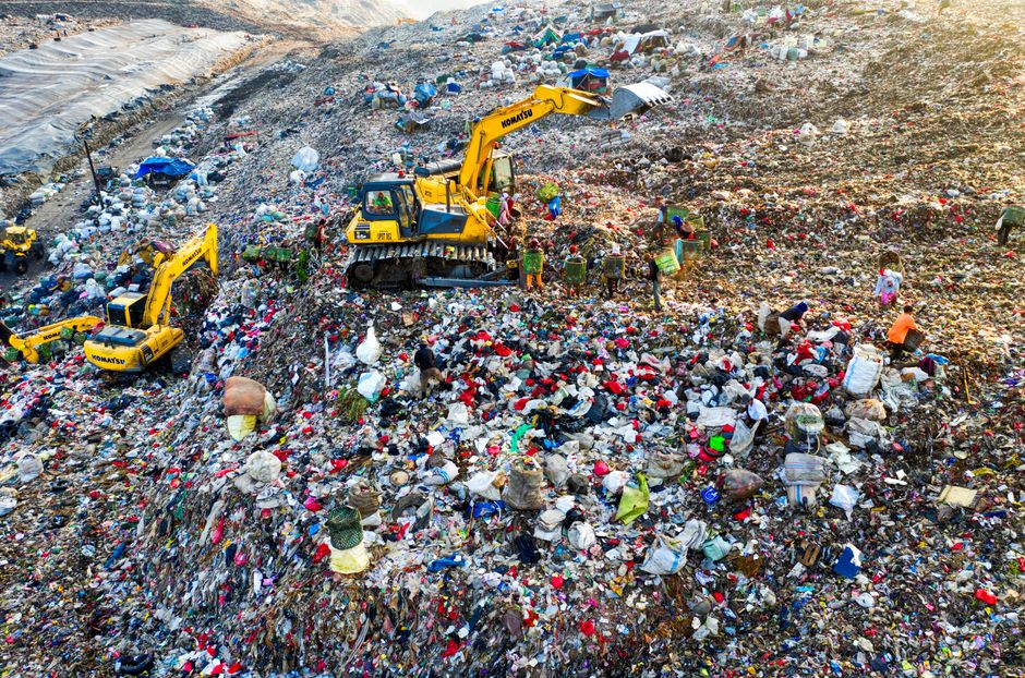 A large landfill site filled with a vast accumulation of mixed waste materials, including plastic bags, paper, textiles, and other household refuse, spread across uneven terrain. In the foreground, several yellow Komatsu excavators are actively engaged in managing the waste, with their mechanical arms extended, lifting and moving debris. The ground surrounding the machinery is heavily layered with rubbish, creating a chaotic and cluttered landscape. Scattered piles of garbage extend into the background, with some areas showing signs of being excavated or pushed aside for disposal. The environment appears dry and dusty, with dirt and gravel mixed into the waste. There are a few workers or operators visible near the machinery, dressed in protective clothing. This scene exemplifies the challenges of waste management and the role of heavy equipment in landfill operations, aligning with alternative waste handling methods outside of local authority collections, as managed by waste removal services such as Rubbish Clearance Croydon.