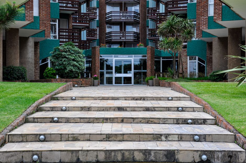Front view of a modern residential or commercial building with a symmetrical facade, featuring multiple balconies with dark wooden railings and teal panels. In the foreground, there is a broad stone staircase with seven steps leading up to the entrance, each illuminated by small, round outdoor lighting fixtures placed evenly along the edges. The staircase is flanked by neatly maintained green grass lawns, separated from the steps by red brick borders. To the left of the stairs, a large, lush shrub with dark green leaves is visible, while to the right, a tall palm-like plant with multiple slender trunks and clusters of long, narrow leaves adds tropical greenery. The entrance comprises large glass doors and windows under a metal canopy, with the sign “EUROMAR” above the doors. The overall scene is well-lit with natural daylight, and the setting appears clean and landscaped, consistent with a private apartment complex or hotel exterior, suitable for independent estate or property clearance services including rubbish removal and site clearance by companies like Rubbish Clearance Croydon.