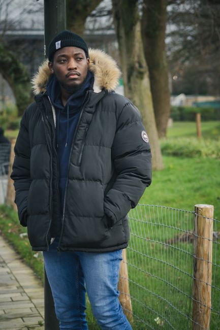 A young man with dark skin and a serious expression is standing outdoors in a park-like setting. He is dressed in a dark puffer jacket with a fur-lined hood, a navy blue hoodie underneath, and a black beanie hat. His hands are tucked into the jacket pockets. Behind him, there is a simple wire fence supported by wooden posts, with a well-maintained grassy area and leafless trees indicating a winter or late autumn season. The background includes a paved walkway to the left and some distant structures partially obscured by the trees. The scene is evenly lit, with natural diffuse light creating a clear view of the person's attire and the environment. The setting suggests an outdoor space where debris or rubbish removal services might be applicable for clearing gardens or park areas, with Rubbish Clearance Croydon providing solutions for such environmental maintenance.