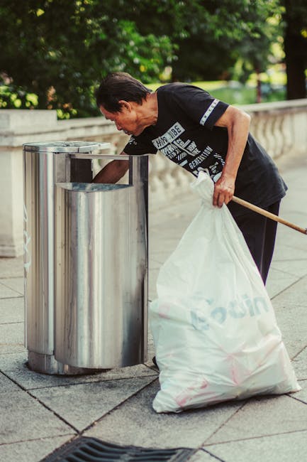A middle-aged woman with short dark hair and light skin is seen emptying a white trash bag into a cylindrical, stainless steel public waste bin situated on a paved sidewalk. She is wearing a black T-shirt with white and grey text, and is holding a wooden broomstick or pole in her left hand. In the background, there is a stone balustrade and lush green trees, suggesting an outdoor park or urban public space. The scene is illuminated by natural daylight, with soft shadows cast on the ground. The stainless steel bin has a reflective, smooth finish, and the white trash bag appears to be made of plastic, filled with waste and secured at the top. This image exemplifies individual efforts in waste disposal, fitting with private or independent rubbish removal activities, possibly carried out by Rubbish Clearance Croydon for environmental cleanliness or targeted rubbish collection services.