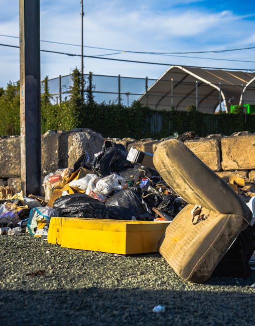 A close-up view of outdoor rubbish piled on a gravel surface, consisting of black and clear plastic bags filled with waste materials, alongside a yellow plastic container and a large, dirty, off-white upholstered seat or cushion which appears worn and stained. The background features a low stone wall made of roughly cut, beige-brown bricks, with some greenery including bushes and trees behind it, and a tall metal pole on the left side of the image. Overhead power lines stretch across a blue sky with scattered white clouds. In the upper right background, part of a semi-open, curved shelter structure is visible, possibly part of a sports or community facility, with a fence and some blurred greenery beyond. The scene suggests an area awaiting waste collection or clearance, relevant to private rubbish removal services such as those offered by Rubbish Clearance Croydon, highlighting the typical materials accumulated outside residential or commercial properties before disposal or recycling.