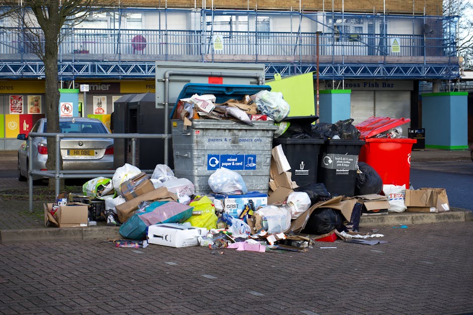 A close-up view of outdoor rubbish piled on a gravel surface, consisting of black and clear plastic bags filled with waste materials, alongside a yellow plastic container and a large, dirty, off-white upholstered seat or cushion which appears worn and stained. The background features a low stone wall made of roughly cut, beige-brown bricks, with some greenery including bushes and trees behind it, and a tall metal pole on the left side of the image. Overhead power lines stretch across a blue sky with scattered white clouds. In the upper right background, part of a semi-open, curved shelter structure is visible, possibly part of a sports or community facility, with a fence and some blurred greenery beyond. The scene suggests an area awaiting waste collection or clearance, relevant to private rubbish removal services such as those offered by Rubbish Clearance Croydon, highlighting the typical materials accumulated outside residential or commercial properties before disposal or recycling.